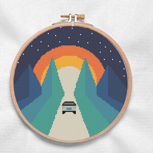 Può includere: Un telaio da ricamo a punto croce con un paesaggio pixelato. Il design presenta un'auto che guida verso un tramonto tra le montagne. Il cielo è blu scuro con stelle bianche e il tramonto è arancione e giallo. Il telaio è in legno.
