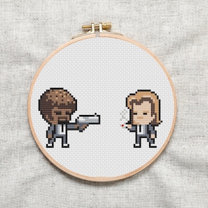 May include: Cross-stitch embroidery hoop with a pixelated image of two characters from the movie Pulp Fiction. The character on the left is holding a gun and the character on the right is holding a cigarette.