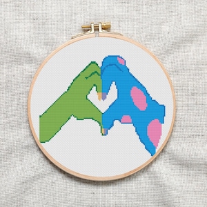 May include: Cross-stitch embroidery hoop featuring a design of two hands forming a heart shape. One hand is green and the other is blue with pink polka dots.