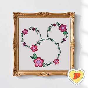 Puede incluir: Arte de punto de cruz en un marco dorado ornamentado. El diseño presenta una forma de cabeza de Mickey Mouse floral, con flores rosas y hojas verdes. El fondo es blanco. Una pegatina amarilla en forma de corazón con la letra "P" está en la esquina inferior derecha.
