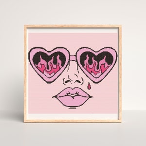 Pode incluir: Um padrão de ponto cruz rosa e preto de um rosto usando óculos de sol em forma de coração com chamas refletidas nas lentes. O rosto também está usando batom rosa e tem uma única lágrima rosa.