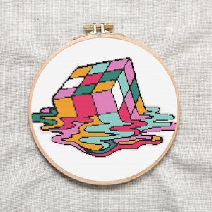 Könnte beinhalten: Ein farbenfroher, pixelig gestalteter Rubik's Cube, der in einer Pfütze von leuchtenden, abstrakten Formen schmilzt. Der Würfel ist eine Mischung aus Rosa, Gelb, Grün und Blau, während die geschmolzenen Formen eine Mischung aus diesen Farben mit zusätzlichem Weiß und Orange sind.