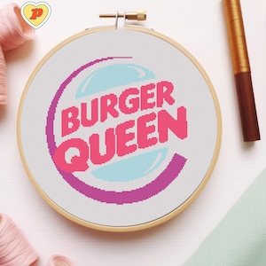 May include: A cross-stitch embroidery hoop featuring the words "BURGER QUEEN" in pink, with a light blue and purple design. The embroidery is set in a wooden hoop, with a gold-colored adjustment screw.