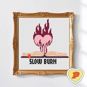 May include: Cross-stitch artwork in a gold ornate frame. The design shows a heart-shaped flame above an open book, with the words "SLOW BURN" below. The heart and flame are pink and red.