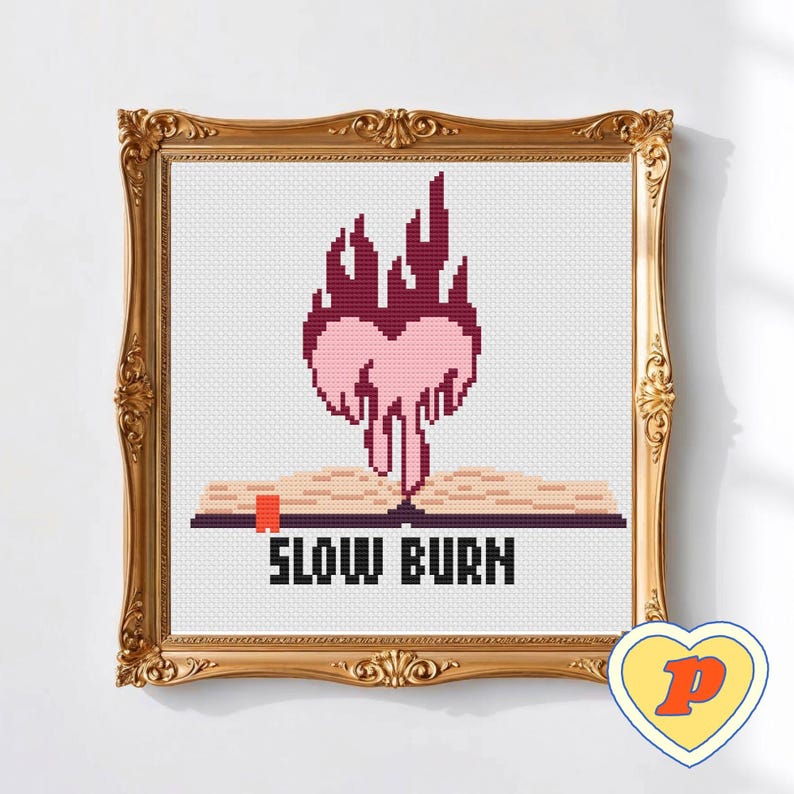 May include: Cross-stitch artwork in a gold ornate frame. The design shows a heart-shaped flame above an open book, with the words "SLOW BURN" below. The heart and flame are pink and red.