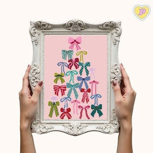 May include: A framed cross-stitch artwork featuring a Christmas tree design made of colorful bows on a pink background. The bows are in shades of pink, green, blue, and red. The frame is white with ornate detailing.