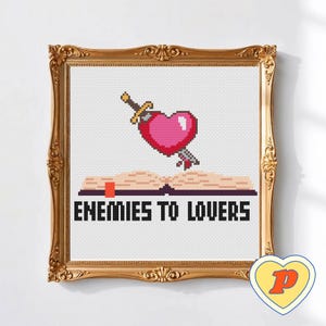 May include: Cross-stitch artwork in a gold ornate frame. The design shows a pink pixelated heart pierced by a sword above an open book. The text "ENEMIES TO LOVERS" is below the book. A small heart-shaped logo is in the bottom right corner.