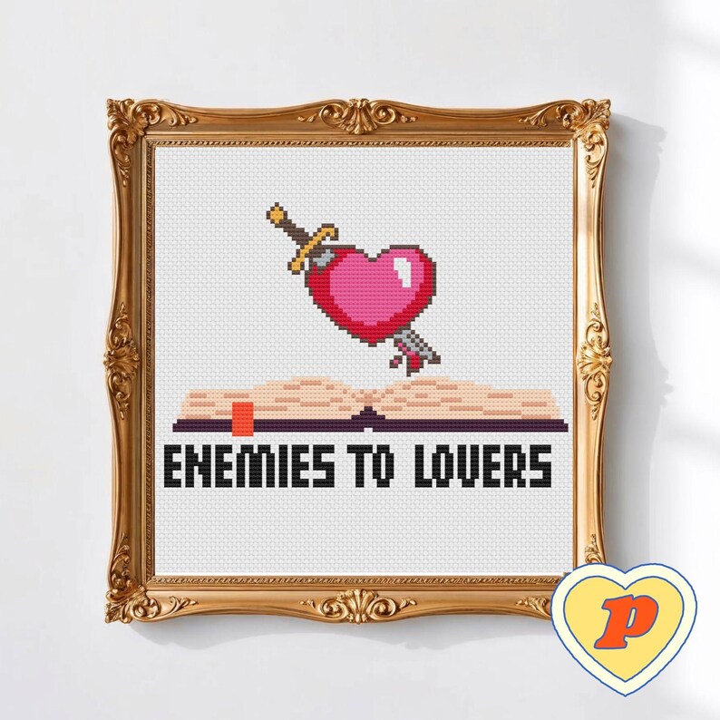May include: Cross-stitch artwork in a gold ornate frame. The design shows a pink pixelated heart pierced by a sword above an open book. The text "ENEMIES TO LOVERS" is below the book. A small heart-shaped logo is in the bottom right corner.