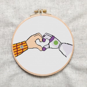 Può includere: Cerchio da ricamo a punto croce con sfondo bianco e due mani di cartoni animati che formano un cuore. Una mano è gialla e arancione con un bottone rosso, l'altra è viola e bianca con un cerchio verde.