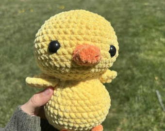 May include: A handmade, yellow crochet chick with black eyes and an orange beak and feet. The plush toy has small wings and a rounded body, perfect for a child's toy or nursery decor.