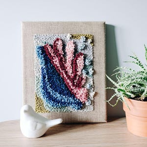 May include: A framed textile art piece with a textured, abstract design. The artwork features a combination of blue, pink, and burgundy yarn, set against a neutral-toned canvas. A small, white ceramic bird sits on a wooden surface.