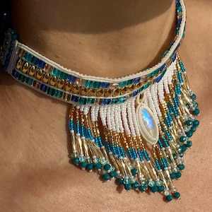 May include: A beaded necklace with a white, gold, teal, and blue color scheme. The necklace features a central pendant and dangling beads. The design incorporates a variety of bead sizes and textures, creating a layered and intricate look. The necklace is worn around the neck.