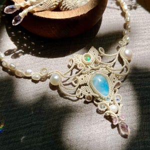 May include: A macrame necklace featuring a central teardrop-shaped blue stone, small white pearls, and iridescent accents. The off-white cord is intricately knotted in a decorative pattern, with a dangling crystal bead at the bottom point.
