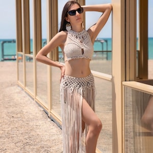 Könnte beinhalten: Beige Makramee-Haltertop und Rock-Set mit Fransen. Das Oberteil hat ein Schlüsselloch-Design, der Rock lange Fransen. Das Set wird an einem Strand mit blauem Ozean im Hintergrund getragen.