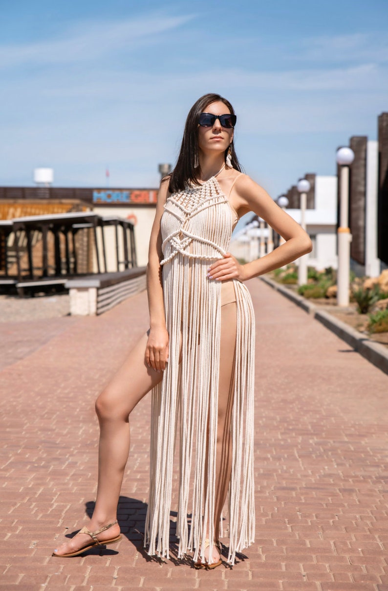May include: A woman wearing a white crochet maxi dress with a fringe hem and black sunglasses. She is standing on a brick pathway.