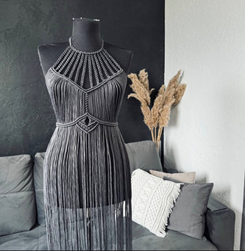 May include: A grey macrame halter top with fringe detailing. The top is displayed on a mannequin against a black background.