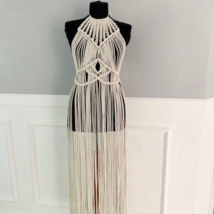 May include: A white crochet halter top with a geometric design and long fringe. The top is displayed on a mannequin.