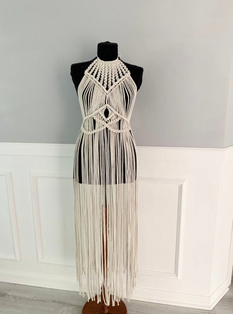 May include: A white crochet halter top with a geometric design and long fringe. The top is displayed on a mannequin.