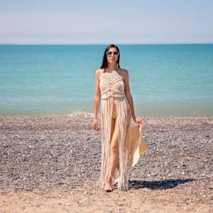 May include: A woman wearing a white crochet maxi dress walks on a beach with a straw hat in her hand. The beach is made of small pebbles and the sea is in the background.