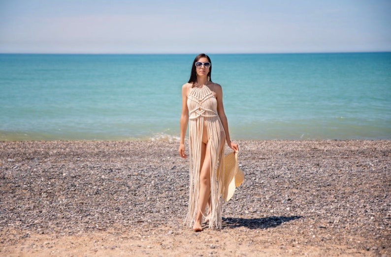 May include: A woman wearing a white crochet maxi dress walks on a beach with a straw hat in her hand. The beach is made of small pebbles and the sea is in the background.