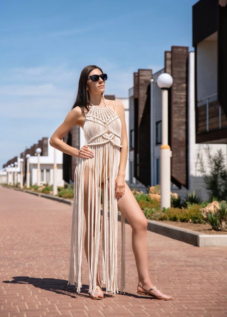 May include: A woman wearing a white crocheted maxi dress with a fringe hem. She is standing on a brick pathway in front of a row of buildings.