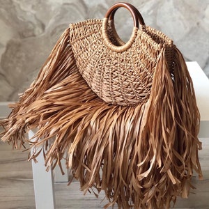 Puede incluir: Bolso de mano de paja tejida con forma redonda y asas de madera. El bolso presenta largos bordes con flecos en color marrón claro, creando un estilo bohemio. Diseñado para llevar objetos, es adecuado para diversas ocasiones.