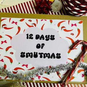 May include: A festive card with the text "12 Days of Smutmas" in a cloud-shaped design. The card is decorated with chili peppers wearing Santa hats and pink bows. A candy cane and silver tinsel add to the holiday theme.