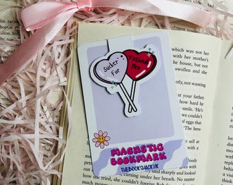 May include: A purple and white bookmark with a pink and white heart lollipop with the text "Sucker For" and a red and white heart lollipop with the text "Fictional Men". The bookmark also has a yellow smiley face with a pink outline and the text "MAGNSTIC BOOKMARK THEBOOKSHACK.UK".