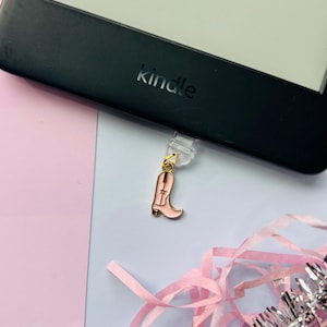 May include: A pink and gold cowboy boot charm attached to a black Kindle e-reader. The charm is hanging from a clear plastic clip. Pink and silver decorative ribbons are in the foreground, with a pink and blue background.