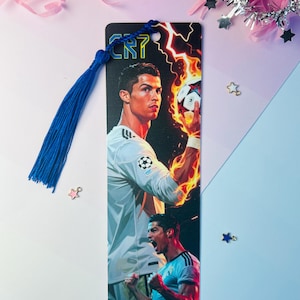 Könnte beinhalten: Rechteckiger Lesezeichen mit einem Porträt von Cristiano Ronaldo und dem Text "CR7". Das Lesezeichen hat eine blaue Quaste und ein Design von Ronaldo, der einen Fußball mit Flammen hält. Der Hintergrund ist ein Farbverlauf aus Blau und Rosa.
