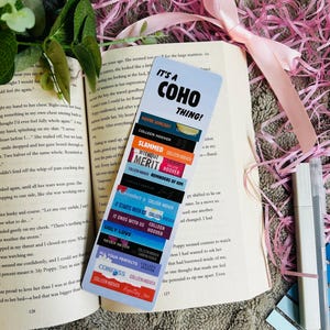 Könnte beinhalten: Nahaufnahme eines Lesezeichens mit blauem Hintergrund und weißem Text, der "It's a Coho Thing!" lautet. Das Lesezeichen besteht aus mehreren kleineren Lesezeichen, die übereinander gestapelt sind, jedes mit einem anderen Buchtitel und Autor. Die Titel umfassen "Maybe Someday", "Slammed", "Without Merit", "Reminders of Him", "November 9", "It Starts With Us", "It Ends With Us", "Ugly Love", "Never Never", "All Your Perfects", "Confess" und "Regretting You".