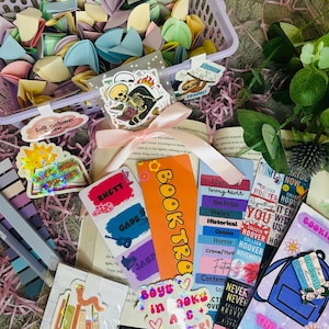 Peut inclure: Une collection de marque-pages, d'autocollants et de papier origami colorés dans un panier violet. Les marque-pages présentent divers motifs et textes, dont "Bookworm", "Boys in Books Are Better" et "The Book Shack."