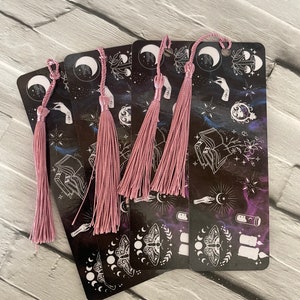 May include: Five black bookmarks with pink tassels. Each bookmark features a different design with a moon, stars, and other celestial imagery. The bookmarks are made of sturdy cardstock and are perfect for marking your place in a book.