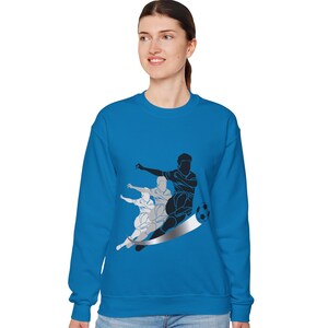 May include: A bright blue sweatshirt featuring a graphic of a soccer player kicking a ball. The graphic is in black and silver, with a trail effect. The sweatshirt is a crew neck style.