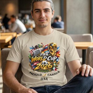 May include: Beige t-shirt featuring a colorful graphic for the FIFA World Cup 2026, with the text "FIFA World Cup 2026" and the flags of Mexico, Canada, and USA. The shirt is worn by a person.