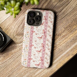 Op de afbeelding: Een smartphonehoesje met een witte achtergrond en een herhalend patroon van roze vlinders, delicate bloemtakjes en verticale stroken van gedetailleerd roze kant. Het hoesje heeft een uitsparing voor een camerasysteem met drie lenzen.