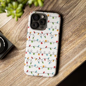 May include: White phone case with a repeating pattern of colorful Christmas lights. The lights are red, green, blue, and yellow, strung on black wires. The case is on a wooden surface.