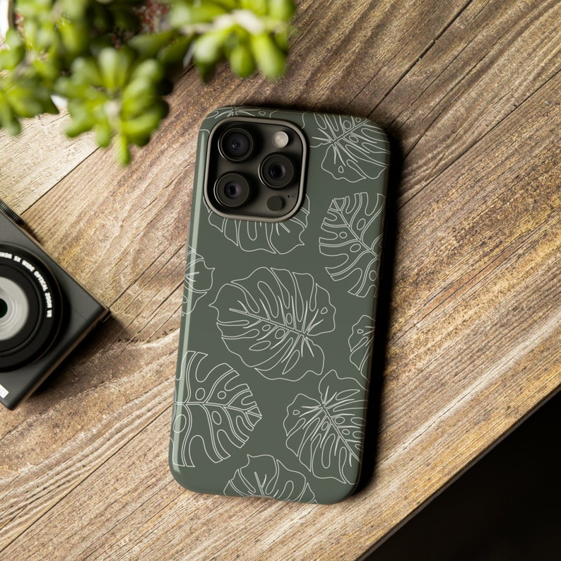 May include: A green phone case with a white outline of tropical leaves. The case has a camera lens cutout.
