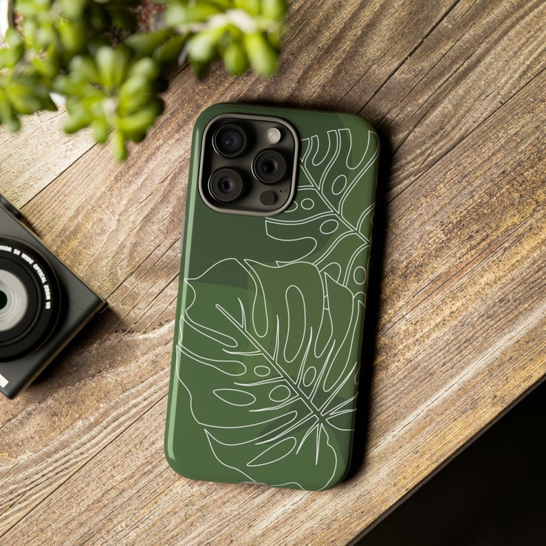 May include: A green phone case with a white outline of tropical leaves. The case has a camera lens cutout and a raised lip around the edges.