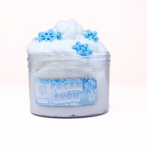 May include: A clear plastic jar filled with white slime, topped with white foam and blue snowflake decorations. The jar has a label that reads "FRESH SNOW" in blue text with snowflake graphics. The background is a soft pink.