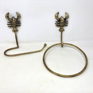 May include: Two gold-toned metal lobster-shaped towel hooks with a ring for hanging towels. The hooks are mounted on a white wall.