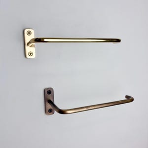 May include: Two metal towel racks. The top rack is a light gold color, and the bottom rack is a darker bronze color. Each rack has a rectangular mounting plate with two screws and a curved bar for hanging towels. The racks are mounted on a light gray wall.