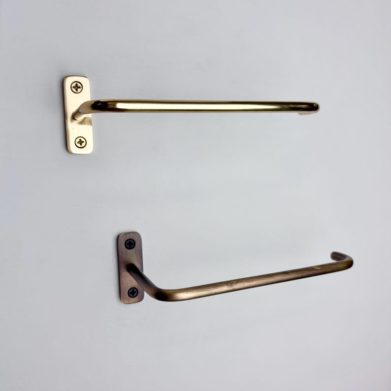 May include: Two metal towel racks. The top rack is a light gold color, and the bottom rack is a darker bronze color. Each rack has a rectangular mounting plate with two screws and a curved bar for hanging towels. The racks are mounted on a light gray wall.