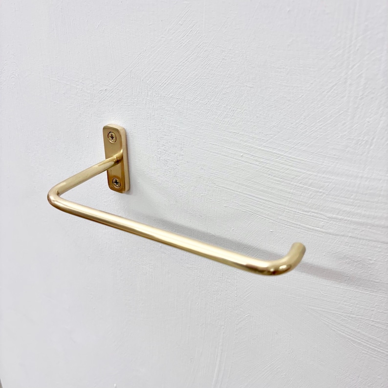 May include: A gold-colored, wall-mounted toilet paper holder. The holder has a rectangular base with two screws and a curved bar for holding the toilet paper roll. The holder is mounted on a white textured wall.