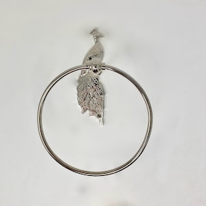 Puede incluir: Un toallero plateado con diseño de pavo real. El pavo real está posado sobre el anillo, con sus detalladas plumas de la cola extendiéndose hacia abajo. El anillo es circular y está hecho de metal brillante.