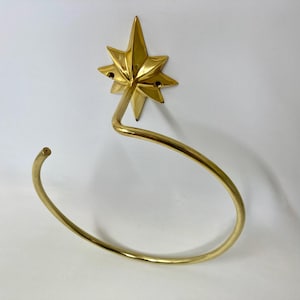May include: A gold-colored metal towel ring with a starburst design. The starburst is mounted on a curved arm that forms the ring. The ring is open at one end.