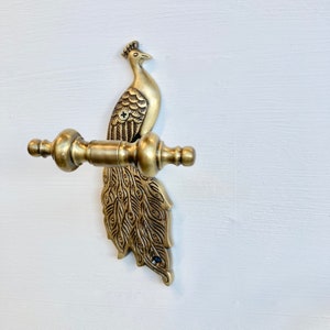 May include: A gold-toned peacock-shaped towel bar with intricate details. The bar is mounted on a white wall.