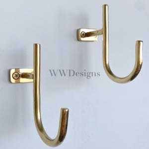 May include: Two polished brass wall hooks with a J-shaped design, each featuring a rectangular mounting plate secured by a single visible screw. The text WWDesigns is centered between the two metallic hooks against a plain white background.
