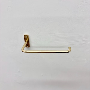 May include: A wall-mounted, brass-colored paper towel holder. The holder has a rectangular mounting plate with two screws and a curved bar for holding paper towels. The design is minimalist and modern, with a focus on functionality and style.
