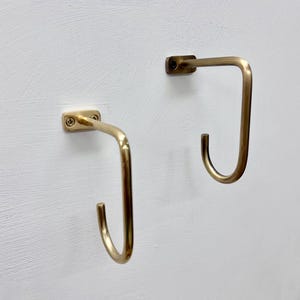 Puede incluir: Dos ganchos de pared de metal color latón. Cada gancho tiene una base rectangular con orificios para tornillos y un gancho curvo en forma de J. Los ganchos están montados en una pared blanca texturizada, ofreciendo un diseño minimalista.
